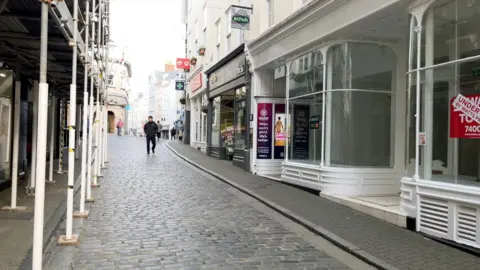 A cobbled street with shops either side and scaffolding outside the buildings on the left side with a few people walking along the street