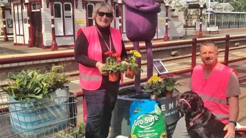 Kettering Civic Society Monica Ozdemir and David Rose, in a railway station. David is to the right and kneeling down with a black dog, by a woman, with jeans on, black top and pink high-vis jacket, they are by plants in pots and Monica is holding two plant pots. A trolley is next to them, also with plants in. 