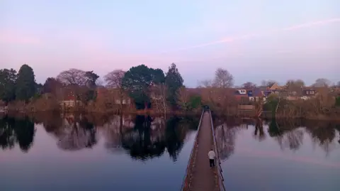 A wooden pedestrian bridge over a body of water. There are trees on the far bank with homes behind. The sky is turning pink.