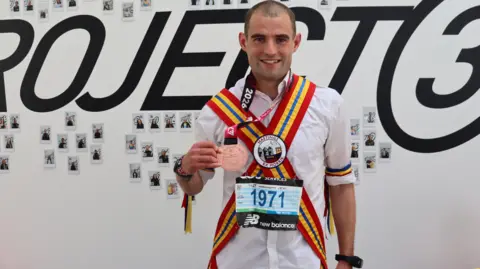Rhis Boorman, wearing his Morris dancing kit of white shirt and white trousers, with two red, yellow and blue striped sashed across his shoulders and chest, proudly shows off his medal to the camera