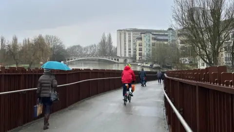 EmmaH A bridge over a river with a brown fence on each side. There is a woman holding a blue umbrella and another person wearing a red coat and a yellow helmet. Over the bridge you can see the river, several high-rise buildings and a second bridge over the river in the distance. 