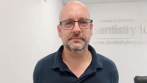 Chris Emmerson is standing in a dental practice. There are white walls behind him. He is wearing a navy top and glasses. 