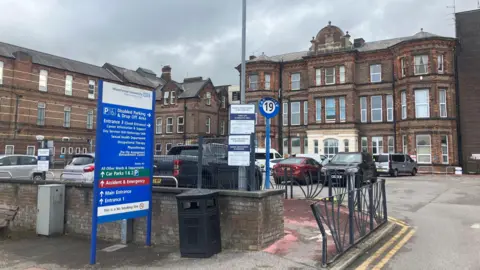 Photograph of North Manchester General Hospital. The image shows the Victorian building, with a surface car park in the foreground.
