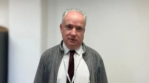 KATE MOSER ANDON/BBC Angus Ellis wearing a grey jacket, white shirt and burgundy tie. He is also wearing a lanyard that says 'councillor'. He is looking at the camera in front of a blurred white background.