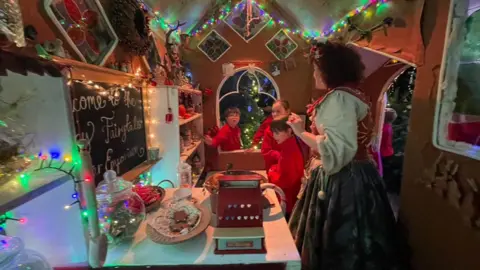 A woman dressed in a Christmas inspired costume, in red and green, is talking to school children who are exploring part of the set. It is made to look like a gingerbread house. There are jars of biscuits on shelves and a red till to serve customers.