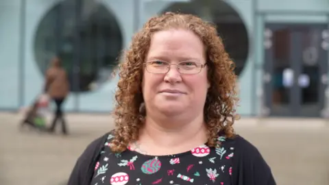 Jamie Niblock/BBC Cheyne Truman is in a dark-coloured top - with floral patterns - and a cardigan. She has curly ginger hair and glasses. She is standing outside facing the camera.