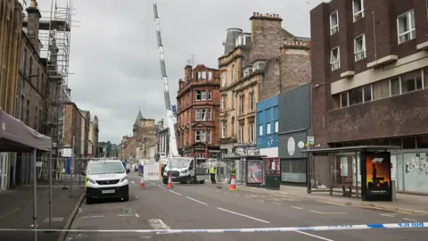 A blocked road with a fire-damaged building in the background. A large crane is visible in the background