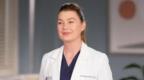 Getty Images Ellen Pompeo in a scene from Grey's Anatomy, dressed as her character Meredith Grey, in navy scrubs and a white doctor's coat. Her blonde hair is pulled back in a ponytail.