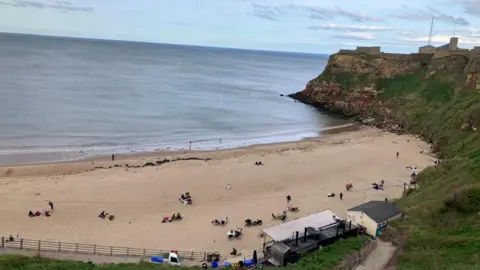 BBC Beach and water on a grey day at Tynemouth. Some people are on the beach and there are cliffs to the right hand side of the cove.