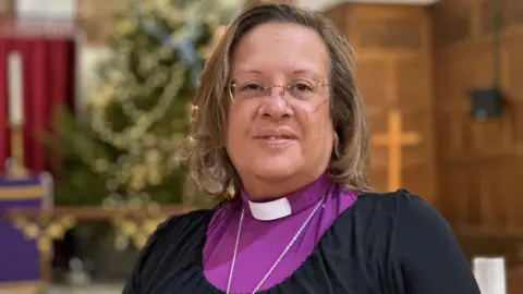 BBC Bishop Tricia Hillas, who has shoulder length brown hair and is wearing glasses, standing in a church. She is wearing a purple clerical shirt with a white clerical collar 