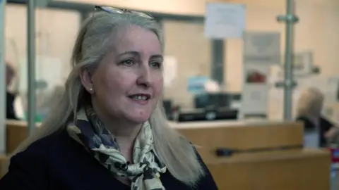 BBC Julie Southern has long grey hair and is wearing a black top and a patterned gold silk scarf. She is stood in a health care reception area.