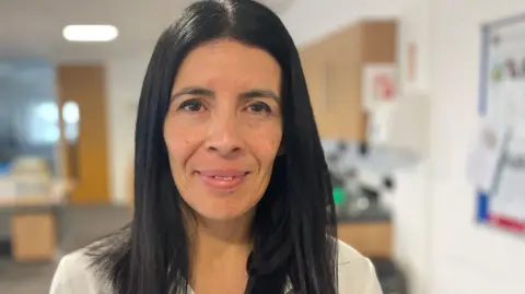 Rocio Cifuentes, who has long dark hair, and is wearing light-coloured clothes, smiles at the camera