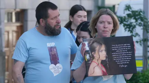 PA Media A man and woman walking next to each other wearing blue t-shirts with 'RIP' written on them with a picture of Kiea. The man has short black hair and beard with tattoos visible on his arm. The woman has short blonde and brown hair as she holds the picture of her daughter alongside a quote.