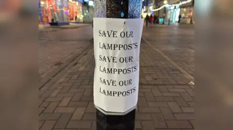 Eastbourne Conservatives The image shows a white sheet reading "Save our lampposts" three times in all capital letters. It is stuck to the neck of a lamppost and a high street can be seen behind it.