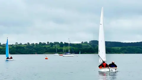 Two men sat in an orange sailing boat on a lake. The man on the right has a blue life jacket, dark clothing and no hair. The man on the left has a red life jacket, a dark coat, green cap and sunglasses. There are other boats and orange buoys on the lake. At the other side of the lake are fields and trees.