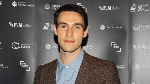 Éanna Hardwicke looking directly at the camera, he has dark hair and is wearing a brown suit with a light blue shirt.