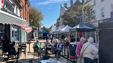 George Carden/BBC People sat outside a cafe in the high street, behind them are gazebos with various market traders in the sunshine
