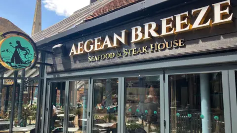 The exterior of Aegean Breeze Seafood & Steak House, featuring large glass doors.