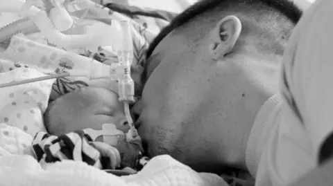 Chestnut A man kissing the cheek of a baby in a hospital bed