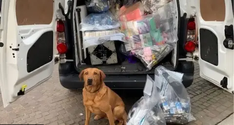 A van has both of its back doors open and there are bags full of illegal vapes. A vape detection dog is also standing next to the vehicle.