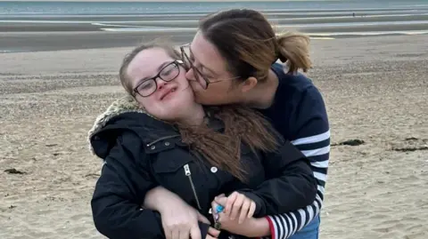 Supplied Emily McAllister, wearing a navy jumper, hugging and kissing her daughter Keira, who has down syndrome and is wearing a black coat, as they stand on a sandy beach. 