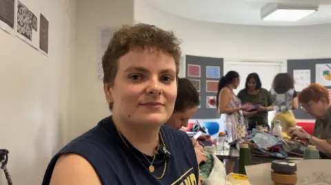 Hannah has short brown hair and is wearing a sleeveless T-Shirt. She is looking towards the camera while sitting in a busy craft room filled with people blurred and crafting in the background.