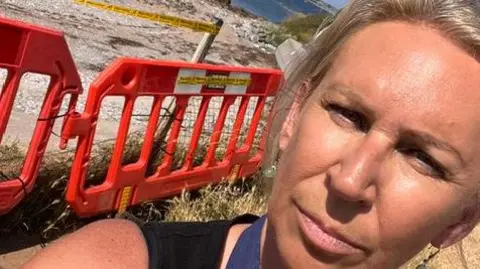 Jane Liew A person has taken a selfie in front of Mount Batten beach, where orange safety barriers and warning tape can be seen blocking off part of the shoreline. In the background, the sea, rocky breakwater and a large ship are visible on a sunny day, highlighting the beach closure.