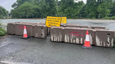 Appleby Town Council Concrete blocks cover the entrance to a car park. There is a yellow road sign which reads 'EDENSIDE CAR PARK CLOSED 01/06 TO 09/06'. There are also two traffic cones in front of the blocks and lots of large trees at the rear of the car park.