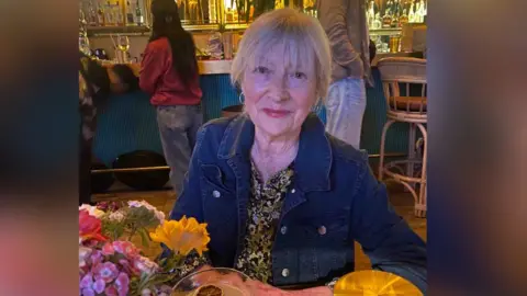 Cathy Kenyon with short blonde/ashy hair sitting at a table wearing a blue denim jacket and a patterned blouse.
