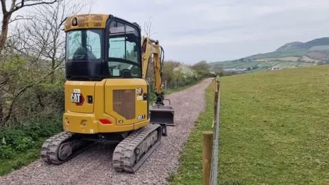 Will Grafton A small digger on a stone track next to a grazing field