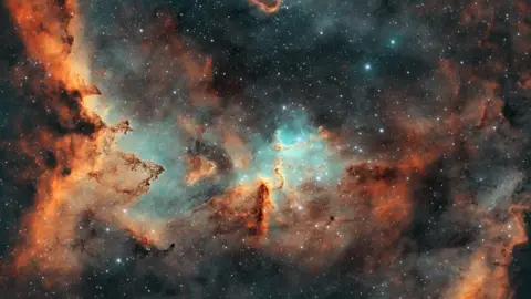 Lee Pullen/UrbanAstrophotography.com The photo shows a vast gas cloud which has spots of light blue and orange in it as well as stars that are twinkling throughout. 