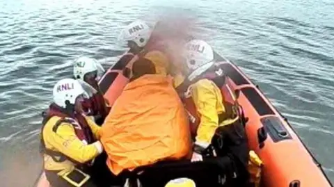 RNLI Queensferry A man in orange blankets on a lifeboat surrounded by four rescuers wearing white helmets and red life jackets.