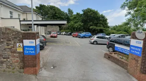 Google The Chadwell Centre in Paignton. The car park outside the white and cream-coloured building is filled with vehicles. A brick wall is on the perimeter of the car park. There are blue and white NHS signs saying the car park is private. Trees are dotted around the opposite side of the car park.