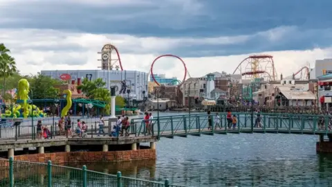 Roberto Machado Noa Orlando Universal Studios with four huge yellow and red rollercoasters in the distance and a lake in the foreground, with a bridge over it. Smaller rides and kiosks in the middle distance, and visitors walking over the bridge.