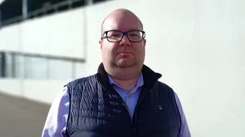 David Brooks is pictured looking into the camera. He's wearing a light coloured shirt with a navy gilet on top. 