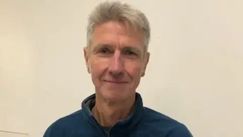 A man with short white hair in a blue zip-up top smiles at the camera. The background behind him is plain.