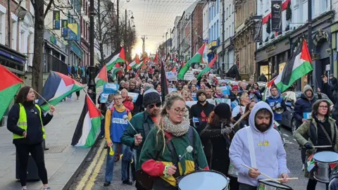 A crowd walking down a street holding Palestinian flags (Green, white, black and red in colour). Some people have drums and signs. Others have PRESS written on their tops.