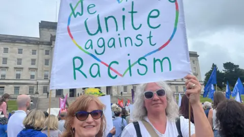 Two women wearing sunglasses hold a banner in front of Stormont that reads 