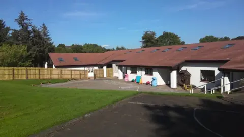 Mike Glenton A single storey school building with a long sloping roof and a white facade. There is a large area of lawn in the front and a wooden fence. 