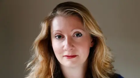 Lexi Ellingsworth A woman with long dark blond hair looks into the camera with a neutral expression. 