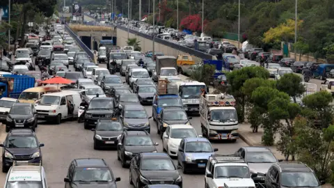 Reuters Traffic jam on a main road in Beirut's southern suburbs, with up to five cars across at one point just as vehicles on a road are joined by those emerging from a tunnel. There are some trees dividing the road, with cars as far as the eye can see on the other side, too.