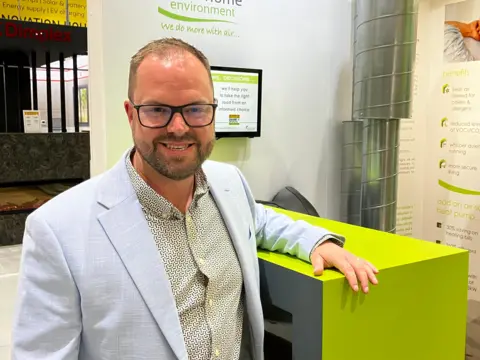 Picture of Harvey Fremlin, managing director of the National Self Build and Renovation Centre wearing a patterned shirt under a light blue jacket. Wearing glasses Harvey's leaning against a heat pump on display at the centre