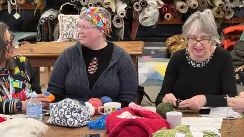 Mousumi Bakshi/BBC Three people are sitting at a table which has knitting equipment on it. Two women on the left and talking to each other and a lady on the right is knitting. Behind them are rolls of fabric on a shelf.