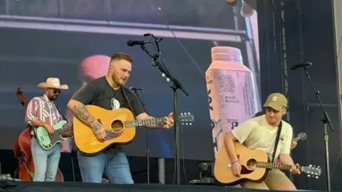 Sarah Sade The photo is of Paul performing with Zach Bryan. They are standing on a stage and are both holding brown acoustic guitars. 