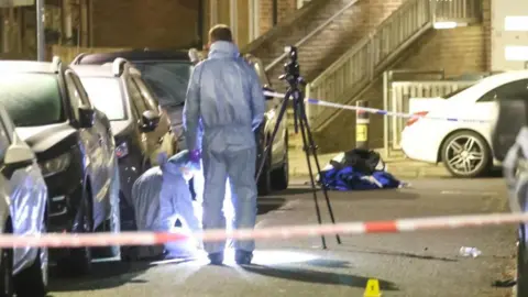 UKNIP A man in a blue forensics suits stands over another person, also in a blue forensics suit, at a crime scene. They are in between lines of parked cars and there is a camera on a tripod next to the man standing. 