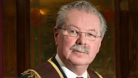 APOD David Hoey is seen wearing glasses, and has short hair and a moustache. 
He is wearing a red and gold collarette over a black suit with a red tie and white shirt. He is standing in front of a wooden placard with writing on it. 