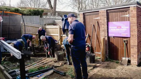 WellChild A group of volunteers in matching dark navy clothing dig and clear ground in a residential back garden on an overcast day. Garden tools, buckets and building materials are scattered across the muddy ground beside a brick outbuilding. A pink sign on the outbuilding reads 'Celebrating 500 WellChild Helping Hands Projects'.