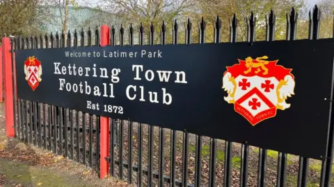 A Kettering Town Football Club sign on a black and red fence.