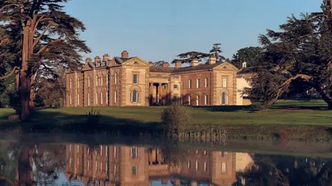 A photograph of the exterior of the Compton Verney manor with trees and grass surrounding it. There is water in the foreground of the image.