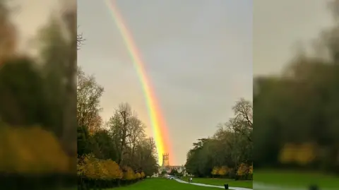 Cirencester Park, Bathurst Estate and Colin Ricalton A bright rainbow shoots down the sky above Cirencester Park, which is surrounded by trees. At the bottom of a long path down the lawn lies St John Baptist Cirencester church, where the rainbow ends.
